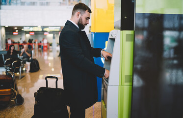 Side view of Caucasian businessman dressed in formal clothing using ATM machine for getting...