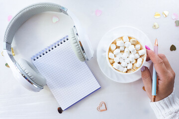 female hands with beautiful manicure hold a cup of coffee and marshmallows and a pen. White headphones with a notepad on a white background with confetti hearts. top view, lifestyle.