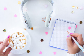 female hands with beautiful manicure hold a cup of coffee and marshmallows and a pen. White headphones with a notepad on a white background with confetti hearts. top view, lifestyle..
