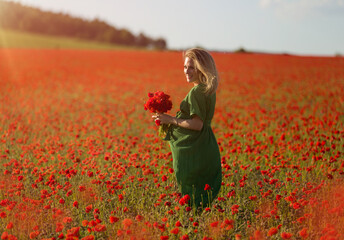 Beautiful blonde pregnant woman in long green dress with a bunch of flowers in hand, in the middle of a red poppy flower field.
