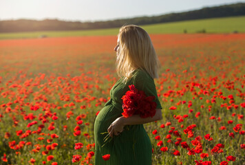 Beautiful blonde pregnant woman in long green dress with a bunch of flowers in hand, in the middle of a red poppy flower field.