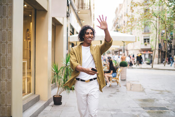Positive ethnic man with smartphone waving hand on street
