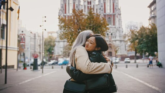 Asian mature mother and adult daughter in casual outerwear are meeting at a city square, smiling, kissing, hugging each other
