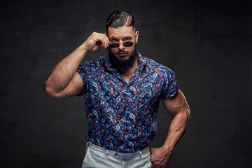 Stylish and powerful guy with huge biceps and with black beard posing in sunglasses in gray background.