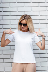 Stylish blonde girl wearing white t-shirt and glasses posing against street