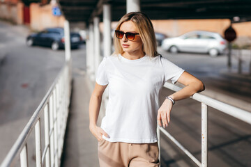 Stylish blonde girl wearing white t-shirt and glasses posing against street