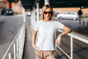 Stylish blonde girl wearing white t-shirt and glasses posing against street