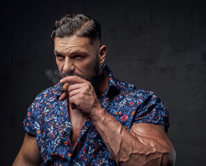Brutal and bearded styled guy in shirt posing and smoking cigar in dark gray background.