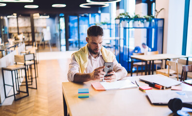 Caucasian male freelancer using mobility connection for checking bitcoin stock exchange during remote job in coworking space, young businessman 30s reading financial news on smartphone website