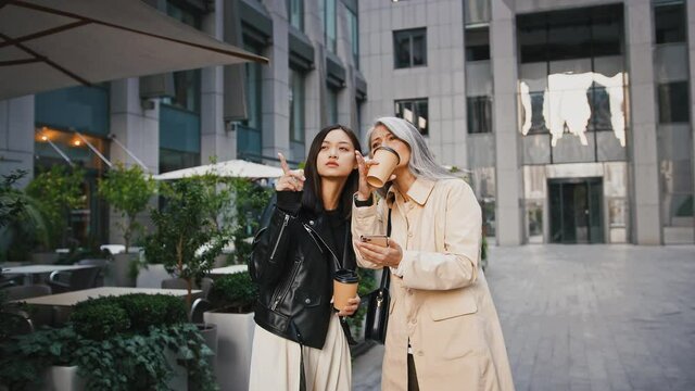 Asian middle-aged female and adult daughter searching for information about buildings around them in smartphone, holding paper cups of coffee