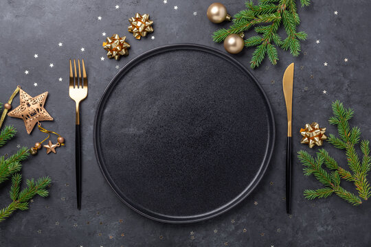 Christmas table setting with empty black ceramic plate, fir tree branch and gold accessories on black stone background. Top view. Copy space