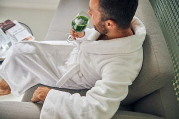 Bearded young man sitting on couch and drinking green smoothie