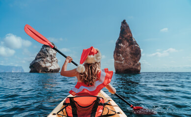 Young brunette woman in red swimsuit and Santa hat, swimming on kayak around basalt rocks like in Iceland. Back view. Christmas and travel concept.