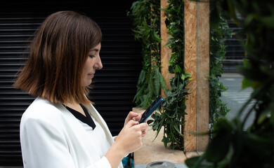 Attractive young female wearing a white suit and drinking coffee from a reusable mug while using her smartphone