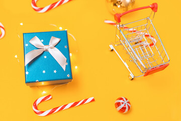 Holiday shopping. A toy shopping cart and a gift box. Yellow background with candys and christmas ball. Close up. Top view. Concept of Christmas sales