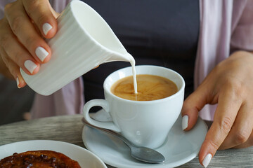 a young woman with beautiful hands and a good manicure sits at a table in a cafe and pours milk into a white cup with hot coffee, next to it lies an appetizing bun with cinnamon, caramel and nuts