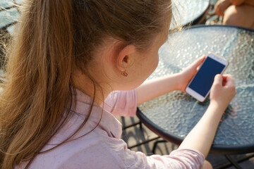 beautiful teenage girl with long blond hair sits on the terrace of a street cafe at a table and uses a smartphone