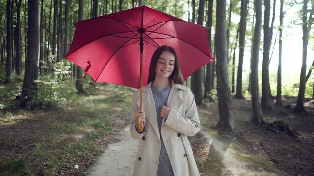 Young brunette woman in casual autumn outfit is strolling along the path in green park forest area, with red umbrella, enjoying beauty of nature, daydreaming, looking happy and thoughtful.