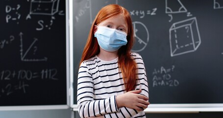 Portrait of pretty small Caucasian red-haired girl with backpack and in medical mask standing in...
