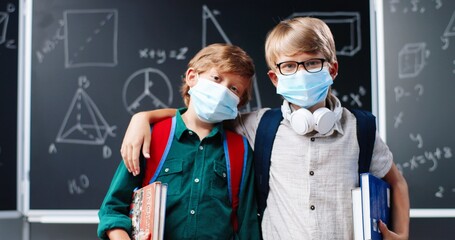 Portrait of cute small Caucasian boys best friends in medical masks, with backpacks and textbooks standing in classroom at blackboard. Little schoolboys hugging in school. Coronavirus pandemic.