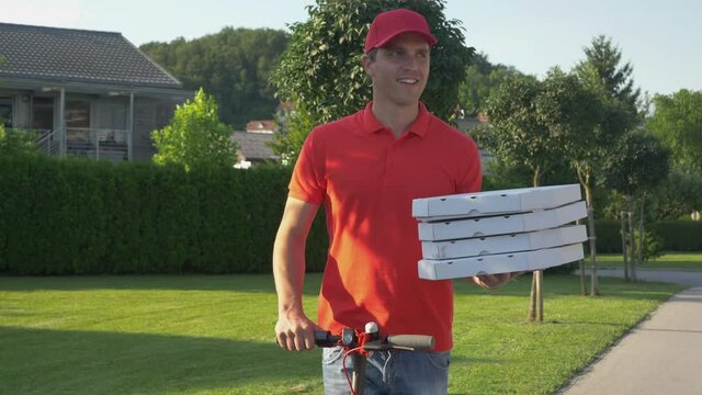CLOSE UP: Cheerful Caucasian courier wearing a red shirt smiles while delivering pizzas in the suburbs on his modern e-scooter. Young pizza delivery guy enjoying a sunny summer day on electric scooter
