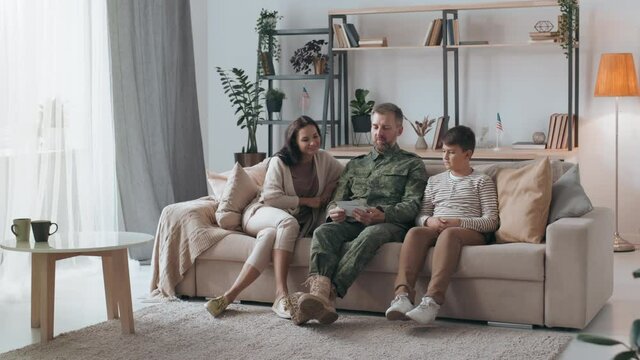 PAN slowmo of male army veteran in military uniform sitting on couch and sharing stories with his family while showing them photos