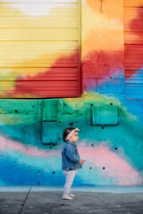 Beautiful toddler girl standing in front of a colorful rainbow mural in the city © Leslie Rodriguez