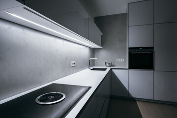 Modern minimal kitchen with white worktop,bright light strip turned on and modern appliances.Focused on hob with integrated extractor hood, then gradually blurs in background(out of focus) direction. - obrazy, fototapety, plakaty