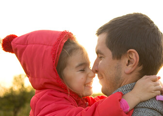 Little girl in red jacket with a hood hugs and kisses her dad, smiles, touches her nose. Happy...