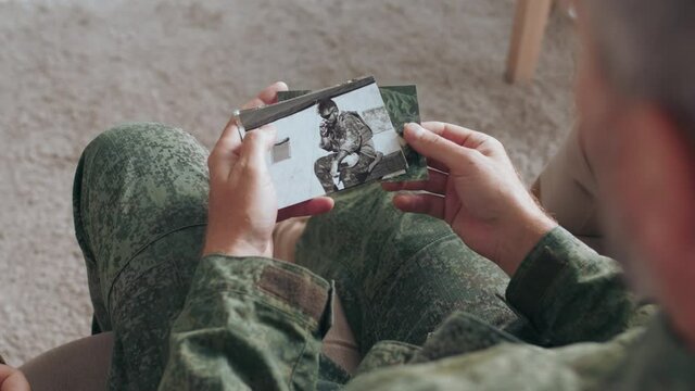 Over the shoulder shot of army soldier or officer in military uniform sitting on couch and showing deployment photos to family while sharing stories