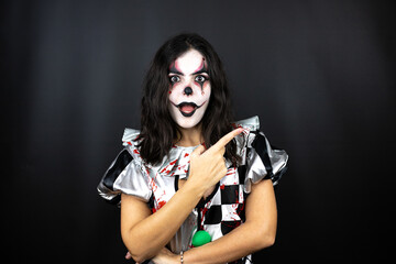 Fototapeta na wymiar woman in a halloween clown costume over isolated black background surprised and pointing with hand and finger to the side