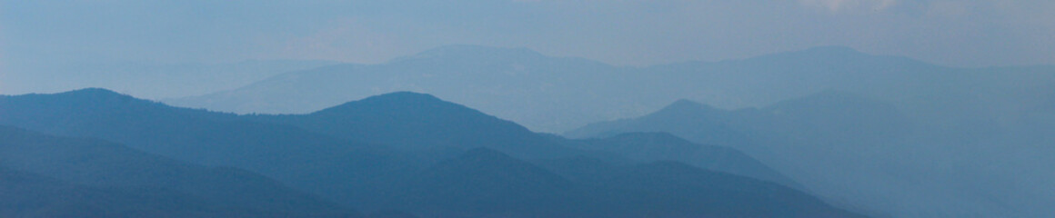 Banner. Blue silhouette of a mountain in the distance, with clouds in the blue sky. Mountains and hills in the distance.