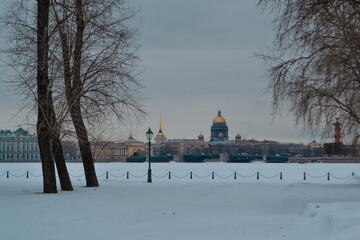 Winter embankment of Saint Petersburg with neat colored buildings and St. Isaac's Cathedral on banks of frozen Neva river in snow with bridge. Foreground park with lantern and bare trees