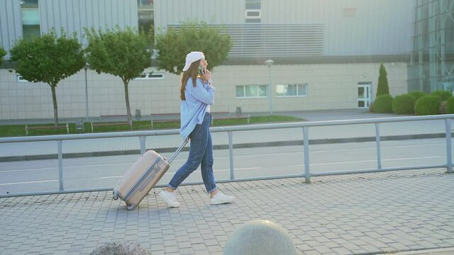 Young Stylish Hipster Girl Tourist Walks With A Suitcase On Wheels And A Phone In Hand Around City On Urban Background A Modern Building. Attractive Female Traveling Woman Student Walking The Street