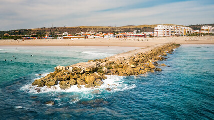 Rocky pier or breakwater on the sandy beach. Beautiful green blue ocean with surfers. Aerial view...