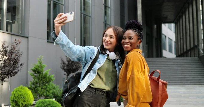 Mixed-races young pretty females smiling cheerfully to camera and taking selfie photo with mobile phone camera at city street. Multi ethnic beautiful happy women students making photos with smartphone