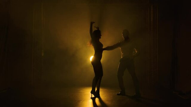 Dancing young couple on a smoky studio background with yellow backlight. Passionate salsa dancers. Silhouette of men and women are moving in slow motion.