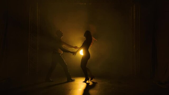 Dancing young couple on a smoky studio background with yellow backlight. Passionate salsa dancers. Silhouette of men and women are moving in slow motion.