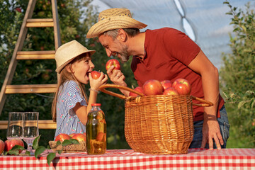 Farmer and His Little Daughter Eating Apples and Having Fun in Sunny Orchard  - Healthy Food Concept