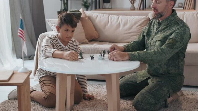 Tilt up shot of bearded male army officer in military uniform and his son kneeling before table in living room and playing with toy soldiers
