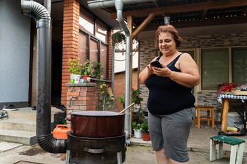 Fototapeta na wymiar Senior woman standing outdoor cooking ajvar or marmalade in her yard at home using mobile phone to check the recipe in summer or autumn day - food preparation concept