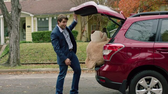 Wide shot of a man opening the trunk of his car and fighting to grab a large teddy bear out of the trunk of his car, falling backwards then walking off camera.
