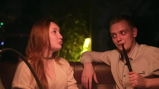 Cute couple in love relaxing on a date. Media. Caucasian young man and woman in black smoking a hookah while sitting at the table in cafe indoors together.