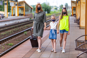 Fototapeta premium Safe family travel. Mother and two daughters in face masks are waiting for take off by train at the railway station. City view.