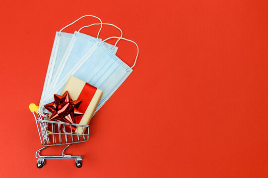 Gift box with a red bow and medical face masks lie in a shopping trolley. Concept of choosing and buying Christmas gifts in quarantine. Christmas sale. Copy space - Powered by Adobe