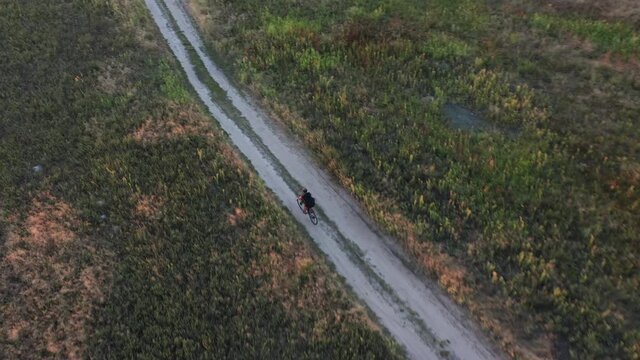 Aerial view of cyclist riding fast along a winding dirt road in a field at golden hour. Drone view. Active lifestyle concept.