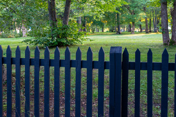 A black wooden picket fence with green grass, large trees and lush shrubs in a garden. The summer scene is vibrant and colorful. The sun is shining on the dark palings giving a warm glow to the wood. 