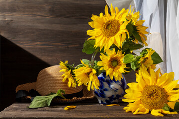 Still life with sunflowers and hat. Bouquet of sunflowers in blue vase and straw hat on an old wooden table in sunlight. Rustic style.