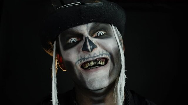 Creepy man face with skeleton makeup in top-hat appearing on black background. Halloween party