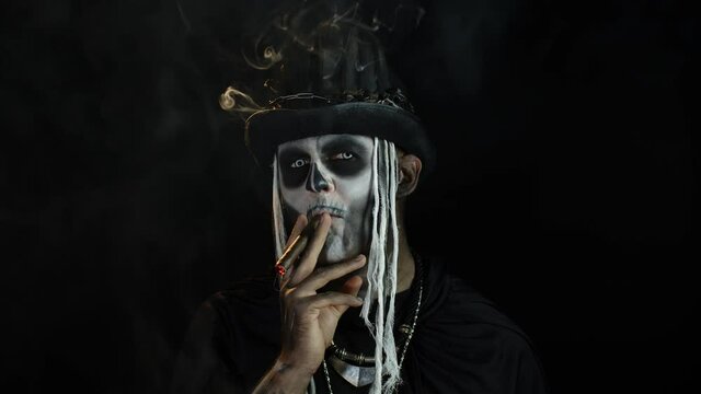 Sinister man with horrible Halloween skeleton makeup smoking cigar, making faces, looking at camera